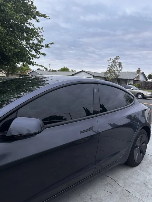Dark grey Tesla Model 3 with tinted side glass, photographed from the side on a residential street